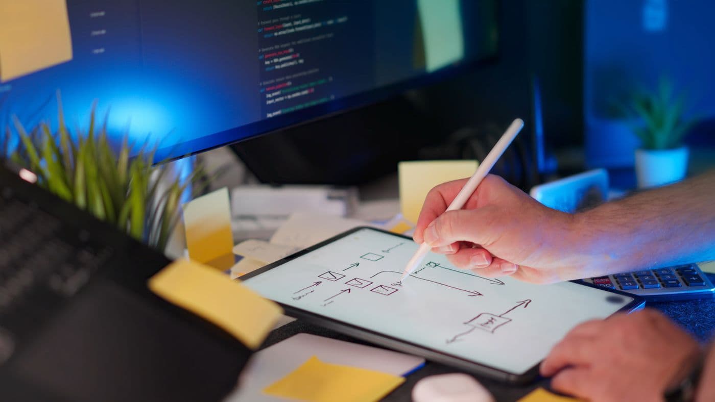 A designer sketching a custom website's user flow on a tablet in front of monitors of code